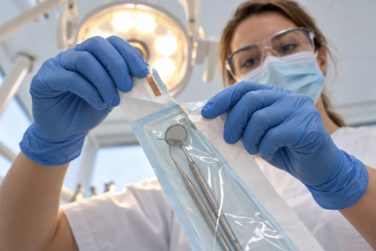 Vue depuis le fauteuil d'un cabinet dentaire : deux mains gantées de bleu ouvrent un sachet de stérilisation contenant un miroir et une sonde dentaires, sous la lampe d'examen, devant un praticien masqué.
