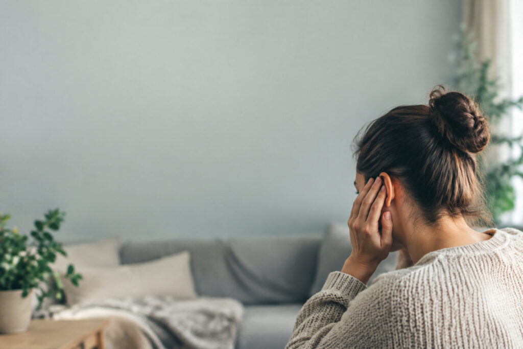 Femme de dos assise dans un salon, touchant son oreille avec une expression de douleur ou d'inconfort.